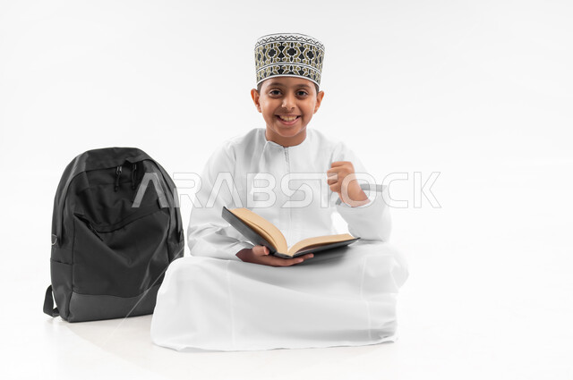 مفهوم الجد والاجتهاد للوصول إلى النجاح، مراجعة الدروس وحل الواجبات، المناهج العلمية التعليمية، بورتريه لطالب عربي خليجي عماني  يرتدي الدشداشة والكمة يجلس بجانب حقيبة الظهر المدرسية يمسك بكتاب يرفع يده بايماءات النصر، صورة لكامل الجسم، خلفية بيضاء