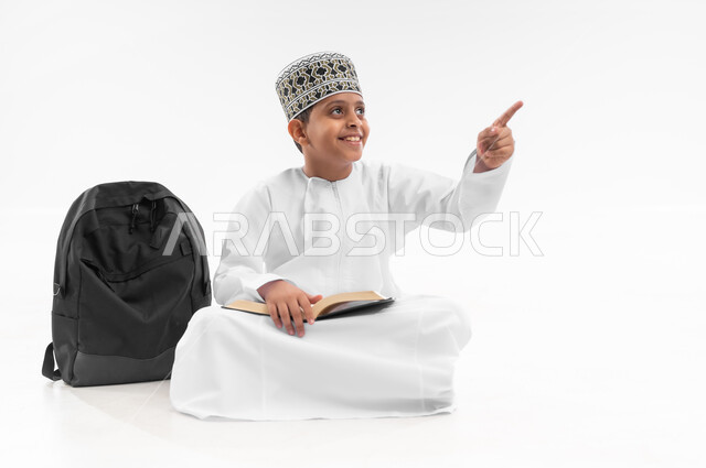 Pointing to a specific location, reviewing lessons and solving homework, educational scientific curricula, portrait of an Arab Gulf Omani student wearing a dishdasha and a kummah sitting next to a school backpack pointing to a place with his hand, full body image, white background