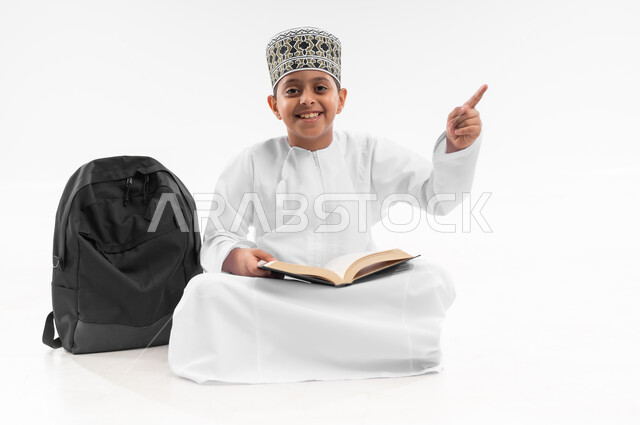 المناهج العلمية التعليمية، الاشارة لموقع معين، مراجعة الدروس وحل الواجبات، بورتريه لطالب عربي خليجي عماني  يرتدي الدشداشة والكمة يجلس بجانب حقيبة الظهر المدرسية يشير بيده لمكان ما، صورة لكامل الجسم، خلفية بيضاء