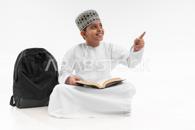 Pointing to a specific location, reviewing lessons and solving homework, educational scientific curricula, portrait of an Arab Gulf Omani student wearing a dishdasha and a kummah sitting next to a school backpack pointing to a place with his hand, full body image, white background