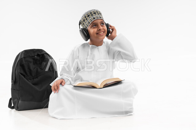 مفهوم الجد والاجتهاد للوصول إلى النجاح، المناهج العلمية التعليمية، بورتريه لطالب عربي خليجي عماني يرتدي الدشداشة والكمة يضع سماعات الرأس يقوم بمتابعة الدروس وحل الواجبات، صورة لكامل الجسم، خلفية بيضاء