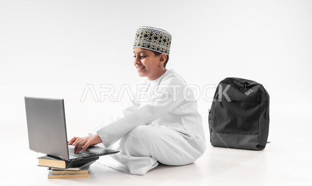 متابعة الحصص و حل الواجبات عبر الانترنت، بورتريه مقرب لصبي عربي خليجي عماني  يرتدي الدشداشة والكمة يقوم بمراجعة دروسه، المذاكرة والدراسة عن بعد، مفهوم التعليم الإلكتروني، استخدام الاجهزة التقنية الحديثة، خلفية بيضاء