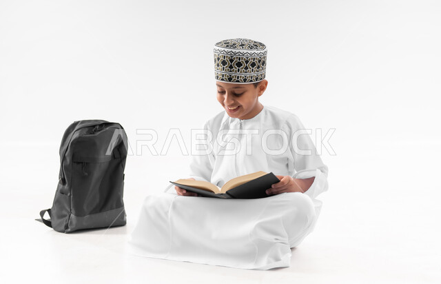 مفهوم الجد والاجتهاد للوصول إلى النجاح، مراجعة الدروس وحل الواجبات، المناهج العلمية التعليمية، العودة الى المدارس، بورتريه لطالب عربي خليجي عماني  يرتدي الدشداشة والكمة يجلس بجانب حقيبة الظهر المدرسية يمسك بيده الكتاب، صورة لكامل الجسم، خلفية بيضاء