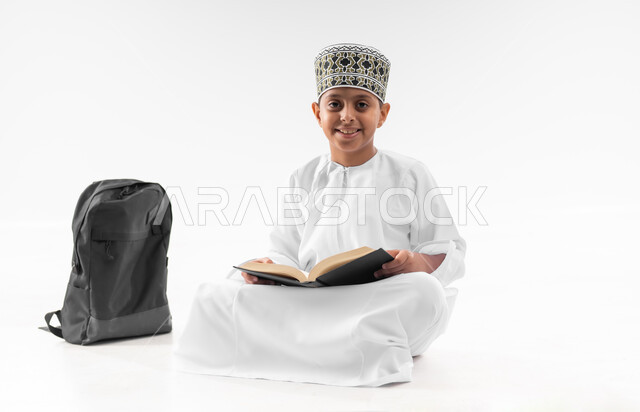 مراجعة الدروس وحل الواجبات، المناهج العلمية التعليمية، العودة الى المدارس، مفهوم الجد والاجتهاد للوصول إلى النجاح، بورتريه لطالب عربي خليجي عماني  يرتدي الدشداشة والكمة يجلس بجانب حقيبة الظهر المدرسية يمسك بيده الكتاب، صورة لكامل الجسم، خلفية بيضاء