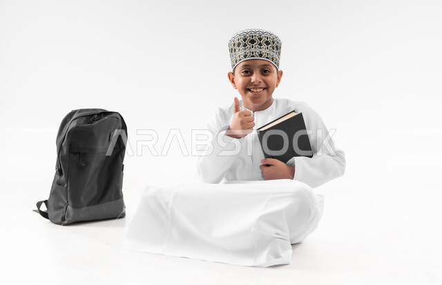 المناهج العلمية التعليمية، العودة الى المدارس، مفهوم الجد والاجتهاد للوصول إلى النجاح، بورتريه لطالب عربي خليجي عماني  يرتدي الدشداشة والكمة يجلس بجانب حقيبة الظهر المدرسية يمسك بيده الكتاب يرفع اصبع الابهام بايماءات الجودة والامتياز، صورة لكامل الجسم، خلفية بيضاء