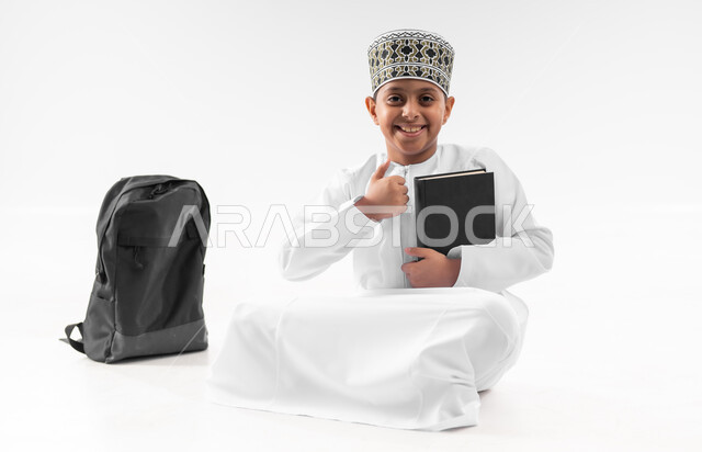العودة الى المدارس، مفهوم الجد والاجتهاد للوصول إلى النجاح، بورتريه لطالب عربي خليجي عماني  يرتدي الدشداشة والكمة يجلس بجانب حقيبة الظهر المدرسية يمسك بيده الكتاب يرفع اصبع الابهام بايماءات الجودة والامتياز، المناهج العلمية التعليمية، صورة لكامل الجسم، خلفية بيضاء