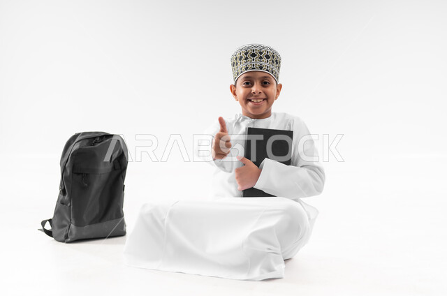 مفهوم الجد والاجتهاد للوصول إلى النجاح، العودة الى المدارس، بورتريه لطالب عربي خليجي عماني  يرتدي الدشداشة والكمة يجلس بجانب حقيبة الظهر المدرسية يمسك بيده الكتاب يرفع اصبع الابهام بايماءات الجودة والامتياز، المناهج العلمية التعليمية، صورة لكامل الجسم، خلفية بيضاء
