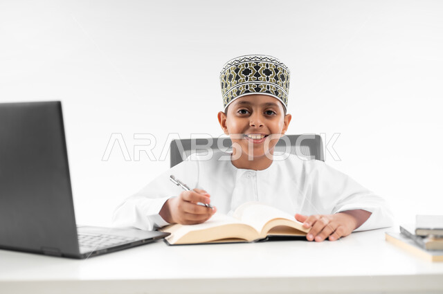 متابعة الحصص و حل الواجبات عبر الانترنت، بورتريه مقرب لصبي عربي خليجي عماني  يرتدي الدشداشة والكمة يقوم بمراجعة دروسه، المذاكرة والدراسة عن بعد، مفهوم التعليم الإلكتروني، استخدام الاجهزة التقنية الحديثة، خلفية بيضاء