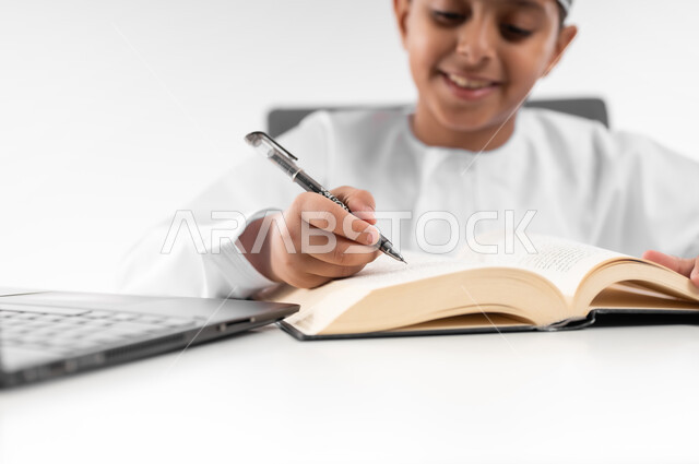 متابعة الحصص و حل الواجبات عبر الانترنت، بورتريه مقرب لصبي عربي خليجي عماني  يرتدي الدشداشة والكمة يقوم بمراجعة دروسه، المذاكرة والدراسة عن بعد، مفهوم التعليم الإلكتروني، استخدام الاجهزة التقنية الحديثة، خلفية بيضاء