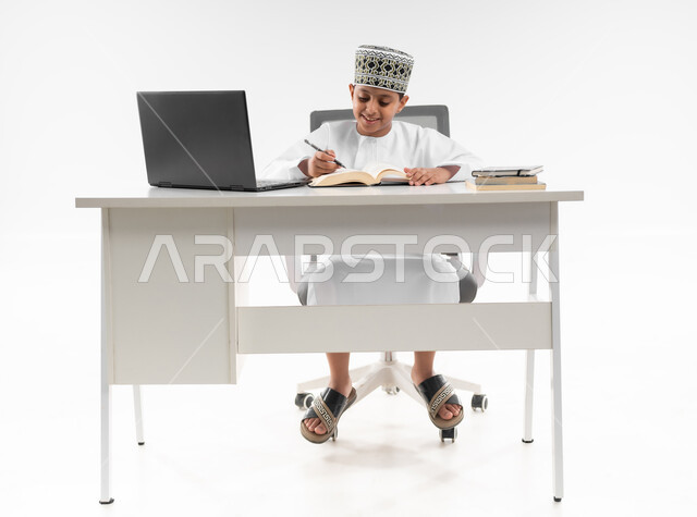 المذاكرة والدراسة عن بعد، استخدام الاجهزة التقنية الحديثة، متابعة الحصص و حل الواجبات عبر الانترنت، بورتريه لصبي عربي خليجي عماني  يرتدي الدشداشة والكمة يقوم بمراجعة دروسه، مفهوم التعليم الإلكتروني، صورة لكامل الجسم، خلفية بيضاء