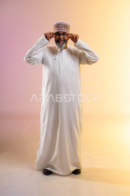 النظر إلى الكاميرا ببشاشة, الشعور بالسعادة والفرح بوضوح الرؤية, استخدام النظارة الطبية لتصحيح البصر, بورتريه لرجل مسن عربي خليجي عماني يرتدي الدشداشه والكمه يضع يديه على نظاراته, مفهوم طول النظر وقصره, صورة الجسم بالطول الكامل, خلفية ملونة