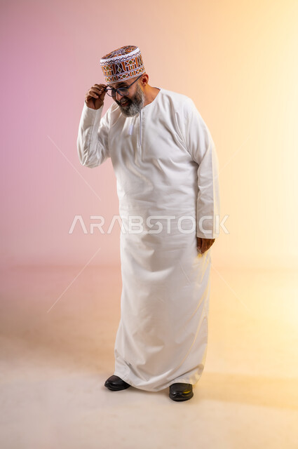 Improving vision and trying to correct vision, contemplating and examining something, the concept of farsightedness and nearsightedness, portrait of an elderly Arab Gulf Omani man wearing a dishdasha and a kimono holding eyeglasses with gestures of concentration, full body image, colored background