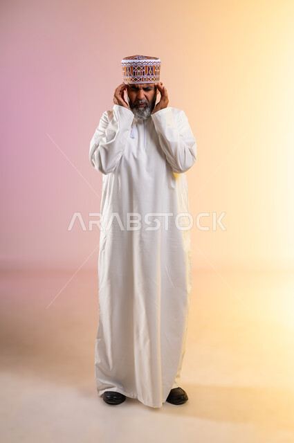 الشعور بالصداع وألم الرأس, الاحساس بالشقيقية والصداع النصفي, الاجهاد والضغط النفسي, بورتريه لرجل مسن عربي خليجي عماني يرتدي الدشداشه والكمه يضع يديه على رأسه ويغمض عينيه بإيماءات الألم الشديد, الإرهاق وفقدان التركيز, صورة الجسم بالطول الكامل, خلفية ملونة
