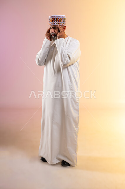 الإرهاق وفقدان التركيز, الشعور بالصداع وألم الرأس, الاحساس بالشقيقية والصداع النصفي, الاجهاد والضغط النفسي, بورتريه لرجل مسن عربي خليجي عماني يرتدي الدشداشه والكمه يضع يديه على رأسه ويغمض عينيه بإيماءات الألم الشديد, صورة الجسم بالطول الكامل, خلفية ملونة