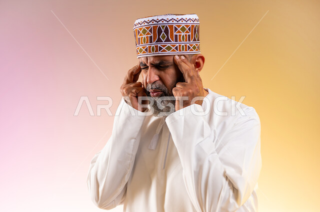 الاحساس بالشقيقية والصداع النصفي, الاجهاد والضغط النفسي, بورتريه مقرب لرجل مسن عربي خليجي عماني يرتدي الدشداشه والكمه يضع يديه على رأسه ويغمض عينيه بإيماءات الألم الشديد, الإرهاق وفقدان التركيز، الشعور بالصداع وألم الرأس, خلفية ملونة