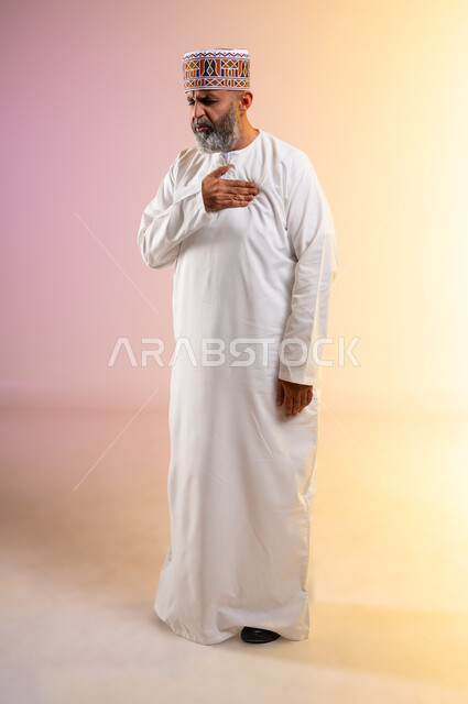 الشعور في زيادة معدل ضربات القلب, الاحساس بذبحه صدريه، بورتريه لرجل مسن عربي خليجي عماني يرتدي الدشداشة والكمه يضع يده على قلبه ويشعر بحالة سيئة من الوخز والألم, اضطرابات وأمراض قلبية, التعب والإرهاق والضغوطات النفسية,  الطول الكامل للجسم، خلفية ملونة