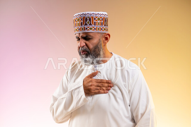 Fatigue, exhaustion and psychological pressure, feeling of increased heart rate, feeling of angina pectoris, close-up portrait of an elderly Arab Gulf Omani man wearing a dishdasha and a sleeve, putting his hand on his heart and feeling a bad state of tingling and pain, heart disorders and diseases, colored background