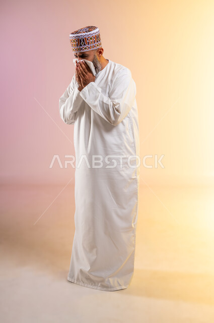 الشعور بالمرض و الزكام والحاجة الى العلاج والدواء, التهاب الأنف التحسسية, بورتريه لرجل مسن عربي خليجي عماني يرتدي الدشداشة والكمه يضع يديه على وجهه بإيماءات العطاس, استخدام المناديل الورقيه, الطول الكامل للجسم، خلفية ملونة