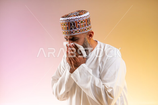 استخدام المناديل الورقيه, الشعور بالمرض و الزكام والحاجة الى العلاج والدواء, التهاب الأنف التحسسية, بورتريه مقرب لرجل مسن عربي خليجي عماني يرتدي الدشداشة والكمه يضع يديه على وجهه بإيماءات العطاس, خلفية ملونة