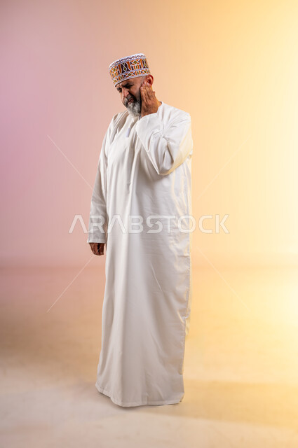 طلب العلاج والمساعدة، تسوس الاضراس و التهاب اللثه، بورتريه لرجل مسن عربي خليجي عماني يرتدي الدشداشة والكمه يضع يده على خده ويغمض عينيه بايماءات الشعور بألم الأسنان، الحاجة لزيارة الطبيب، الجسم بالطول الكامل، خلفية بيضاء