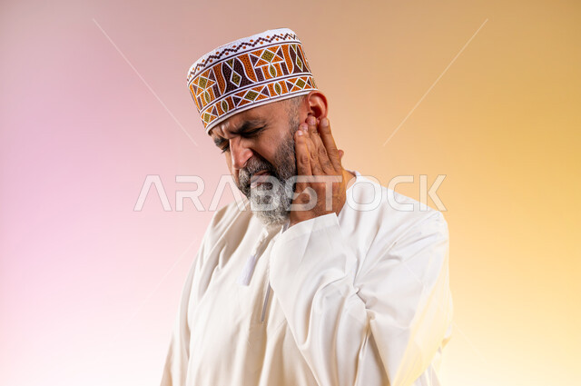 Tooth decay and gingivitis, close-up portrait of an elderly Arab Gulf Omani man wearing a dishdasha and a turban, putting his hand on his cheek and closing his eyes with gestures of toothache, need to visit a doctor, requesting treatment and help, white background