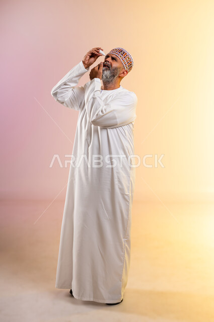 الحاجة للفحص وعلاج حالات التهاب العصب البصري، بورتريه لرجل مسن ضرير عربي خليجي عماني يرتدي الدشداشه والكمه يقوم بوضع قطرة عينية, ارتفاع ضغط العين, الاحساس بالحرقة و احمرار العيون، صورة الجسم بالطول الكامل, خلفية ملونة