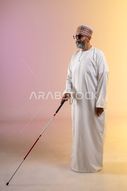 Teaching the blind the techniques of mobility using a cane, portrait of an elderly blind Arab Gulf Omani man wearing a dishdasha and a kuma, wearing sunglasses and carrying a cane, providing psychological support to the blind and enhancing their confidence and motivating them to achieve their potential, full-length body image, colored background