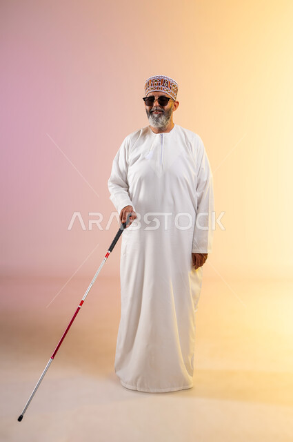 تعليم المكفوفين تقنيات التنقل باستخدام العصا، بورتريه لرجل مسن ضرير عربي خليجي عماني يرتدي الدشداشه والكمه يضع النظارة الشمسية ويحمل العصا، توفير الدعم النفسي للمكفوفين وتعزيز ثقتهم وتحفيزهم على تحقيق إمكانياتهم، صورة الجسم بالطول الكامل, خلفية ملونة