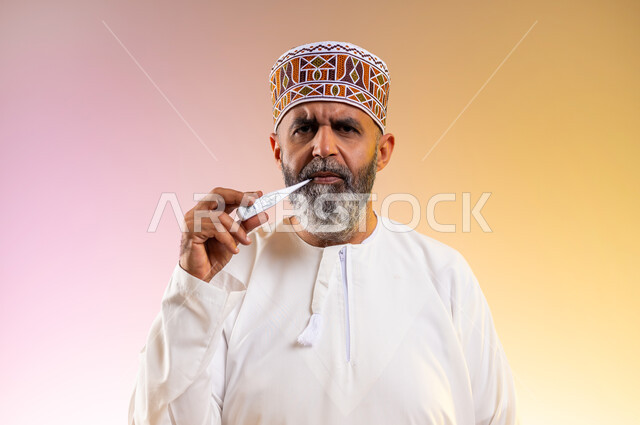 القيام بعرض الميزان الرقمي، ضرورة وجود جهاز مقياس حرارة في المنزل، بورتريه مقرب لرجل مسن عربي خليجي عماني يرتدي الدشداشه والكمه ينظر الى الكاميرا بإيماءات الإرهاق بسبب المرض, الاصابة بالحمى والتعب, خلفية ملونة