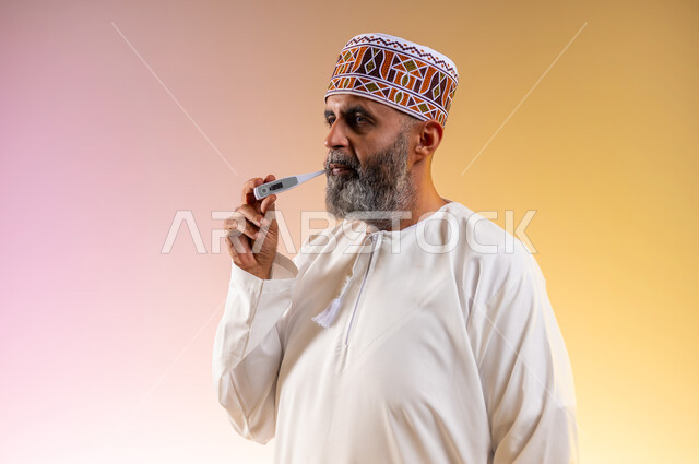 الإحساس بالمرض والإرهاق، بورتريه مقرب لرجل مسن عربي خليجي عماني يرتدي الدشداشه والكمه يقوم بقياس درجة حرارته بإستخدام الميزان الرقمي بتعابير الانزعاج والتعب، ضرورة زيارة الطبيب لوضع الخطة العلاجية، خلفية ملونة