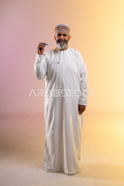 الشعور بالتحسن وانخفاض الحرارة, مفهوم التماثل للشفاء، بورتريه لرجل مسن عربي خليجي عماني يرتدي الدشداشه والكمه يمسك بيده جهاز مقياس الحرارة، النظر الى الكاميرا بتعابير السرور والفرح للتعافي من المرض، صورة الجسم بالطول الكامل, خلفية ملونة