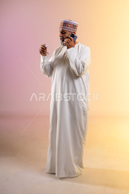 اجراء مكالمة هاتفية عبر الجوال لاستشارة الطبيب عن العلاج المناسب، بورتريه لرجل مسن عربي خليجي عماني يرتدي الدشداشه والكمه يمسك بيده جهاز مقياس الحرارة وينظر الى المؤشر, الحاجة إلى المساعدة الطبية, الشعور بالإرهاق وأعراض الحمى، صورة الجسم بالكامل, خلفية ملونة
