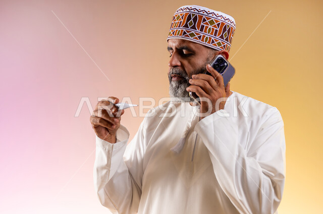 الحاجة إلى المساعدة الطبية, الشعور بالإرهاق وأعراض الحمى، اجراء مكالمة هاتفية عبر الجوال لاستشارة الطبيب عن العلاج المناسب، بورتريه مقرب لرجل مسن عربي خليجي عماني يرتدي الدشداشه والكمه يمسك بيده جهاز مقياس الحرارة وينظر الى المؤشر, خلفية ملونة