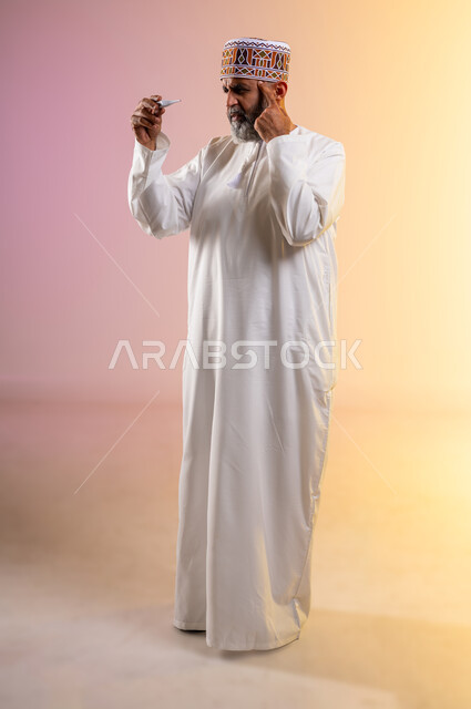 الشعور بالإرهاق وأعراض الحمى، اجراء مكالمة هاتفية عبر الجوال لاستشارة الطبيب عن العلاج المناسب، بورتريه لرجل مسن عربي خليجي عماني يرتدي الدشداشه والكمه يمسك بيده جهاز مقياس الحرارة وينظر الى المؤشر, الحاجة إلى المساعدة الطبية, صورة الجسم بالكامل, خلفية ملونة