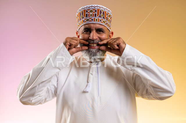Looking at the camera with happy gestures, using porcelain veneers to create a dazzling white smile, importance of dental follow-up, care, cosmetics and whitening, close-up portrait of an elderly Arab Gulf Omani man wearing a dishdasha and a kummah pointing to his teeth, colorful background