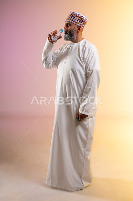 المداومة على شرب المياه، الاكثار من السوائل للحفاظ على الجسد من الجفاف، بورتريه لرجل كبير بالسن عربي خليجي عماني يرتدي الدشداشه والكمه يقوم بشرب المياه العذبة، الجسم بالطول الكامل، المحافظة على سلامة الجهاز المناعي للجسم، خلفية ملونة