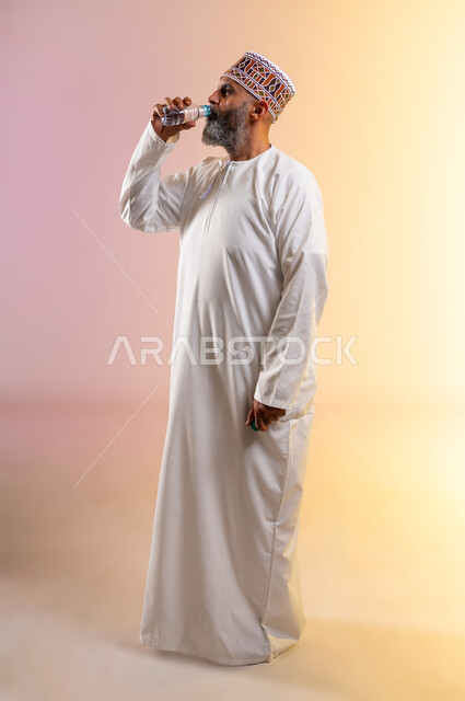 المحافظة على سلامة الجهاز المناعي للجسم، المداومة على شرب المياه، الاكثار من السوائل للحفاظ على الجسد من الجفاف، بورتريه لرجل كبير بالسن عربي خليجي عماني يرتدي الدشداشه والكمه يقوم بشرب المياه العذبة، الجسم بالطول الكامل، خلفية ملونة