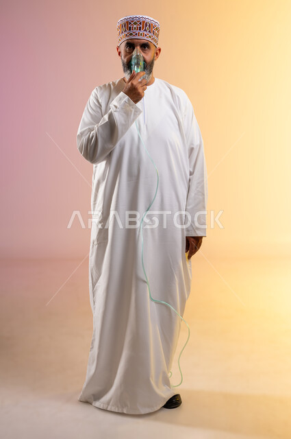 تأمين الراحة للعضلات التنفسية باستخدام جهاز التنفس الصناعي، بورتريه لرجل مسن عربي خليجي عماني يرتدي الدشداشة والكمه يقوم باستنشاق أدوية معالجة لانقطاع التنفس عن طريق قناع الاكسجين، تقديم الخدمات الصحية، معالجة حالات ضيق التنفس، الطول الكامل للجسم، خلفية ملونة