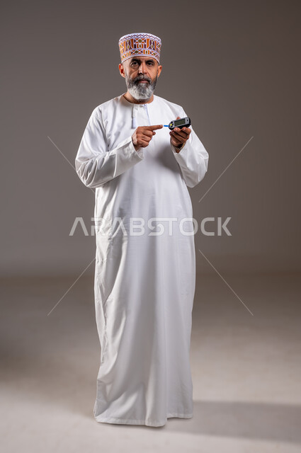 Looking at the camera with tired expression, modern electronic devices for rapid medical examination, full body length, diabetes, portrait of an elderly Arab Gulf Omani man wearing a dishdasha and a kimmah checking blood sugar, gray background