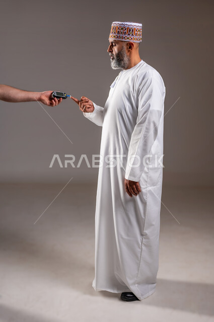 جهاز إلكتروني حديث لمرضى داء السكري, حالات مرضية ومشاكل وآلام جسدية, الرعاية الصحية والفحص الطبي السريع, بورتريه  لرجل مسن عربي خليجي عماني يرتدي الدشداشة والكمة يقوم بفحص نسبة السكر, مشاكل ارتفاع السكر في الدم, الجسم بالطول الكامل، خلفية رمادية