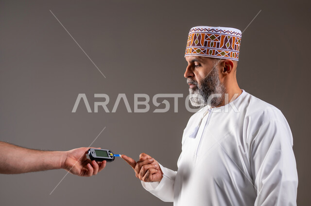 مشاكل ارتفاع السكر في الدم, جهاز إلكتروني حديث لمرضى داء السكري, حالات مرضية ومشاكل وآلام جسدية, الرعاية الصحية والفحص الطبي السريع, بورتريه مقرب لرجل مسن عربي خليجي عماني يرتدي الدشداشة والكمة يقوم بفحص نسبة السكر في الدم,  خلفية رمادية