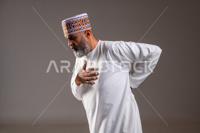 التعب والإرهاق والضغوطات النفسية, الشعور في زيادة معدل ضربات القلب, الاحساس بذبحه صدريه، بورتريه مقرب لرجل مسن عربي سعودي عماني يرتدي الدشداشه والكمه يضع يده على قلبه ويشعر بحالة سيئة من الوخز والألم, اضطرابات وأمراض قلبية, خلفية رمادية