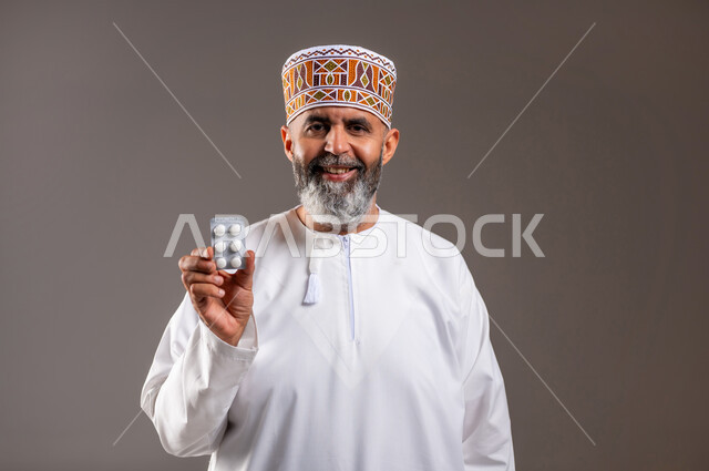 الاهتمام بالصحة والالتزام بالأوقات الصحيحة لأخذ العلاج, استخدام عقار الدواء بوصفة طبية, بورتريه مقرب لرجل كبير بالسن عربي خليجي عماني يرتدي الدشداشه والكمه يقف باستقامة ويحمل شريط الدواء, مفهوم الطب والرعاية الطبية, خلفية رمادية