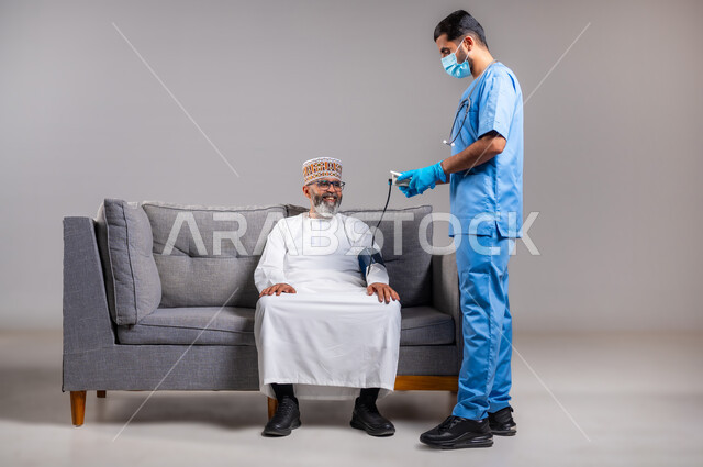العمل في القطاع الصحي، الفحص الطبي والتشخيص السريع والرعاية الصحية المنزلية، بورتريه لممرض يرتدي الكمامة وقفازات الوقاية يقوم بقياس ضغط الدم لمريض عربي خليجي عماني يرتدي الدشداشة والكمه يجلس على الكنبه، متابعة دورية لفحص ضغط، الطول الكامل للجسم، خلفية رمادية