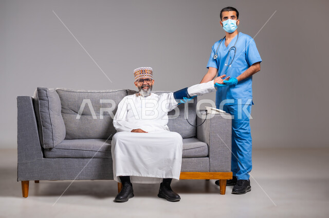 العمل في القطاع الصحي، الفحص الطبي والتشخيص السريع والرعاية الصحية المنزلية، بورتريه لممرض يرتدي الكمامة وقفازات الوقاية يقوم بقياس ضغط الدم لمريض عربي خليجي عماني يرتدي الدشداشة والكمه يجلس على الكنبه، متابعة دورية لفحص ضغط، الطول الكامل للجسم، خلفية رمادية