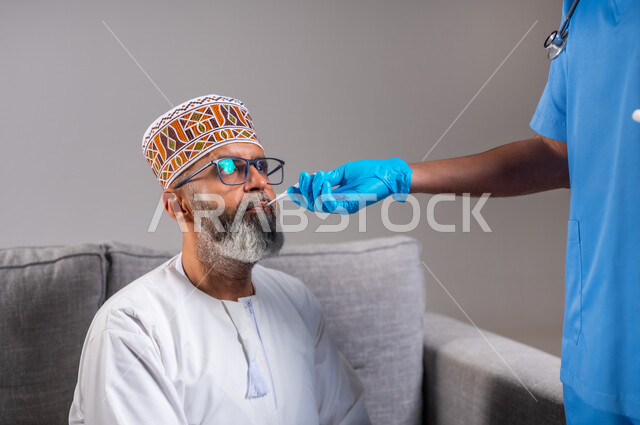 ممرض يلبس الكفوف وكمامة الوقاية يقوم بقياس درجة حرارة الجسم بواسطة جهاز الترمومتر الرقمي، شركات التمريض المنزلي, الحاجة إلى المساعدة الطبية, بورتريه مقرب لرجل مسن عربي خليجي عماني يرتدي الدشداشه والكمه يجلس على الكنبة مصاب بالحمى, خلفية رمادية