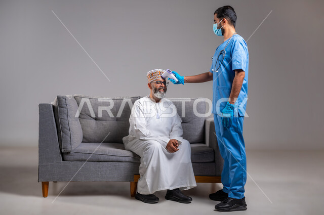 العمل في القطاع الطبي، طبيب يحمل مقياس الحرارة الرقمي بالأشعة تحت الحمراء، إتباع إجراءات الوقاية و السلامة من فايروس كورونا، الوعي الصحي وتجنب إنتشار العدوى، بورتريه لممرض يرتدي اليونيفورم يقوم بفحص مريض مسن عربي خليجي عماني يرتدي الدشداشة والكمه، خلفية رمادية