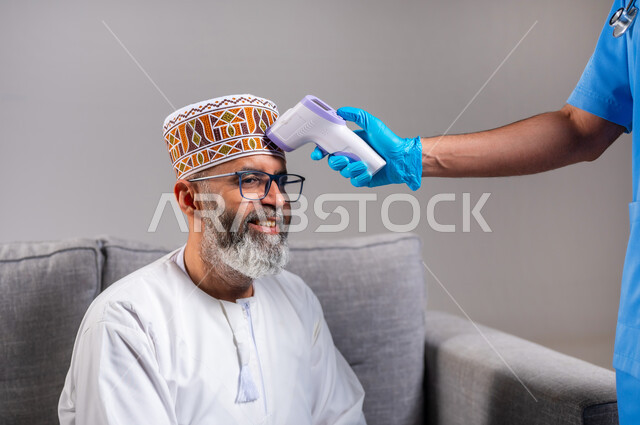 الوعي الصحي وتجنب إنتشار العدوى، بورتريه لممرض يرتدي اليونيفورم يقوم بفحص مريض مسن عربي خليجي عماني يرتدي الدشداشة والكمه، العمل في القطاع الطبي، طبيب يحمل مقياس الحرارة الرقمي بالأشعة تحت الحمراء، إتباع إجراءات الوقاية و السلامة من فايروس كورونا، خلفية رمادية