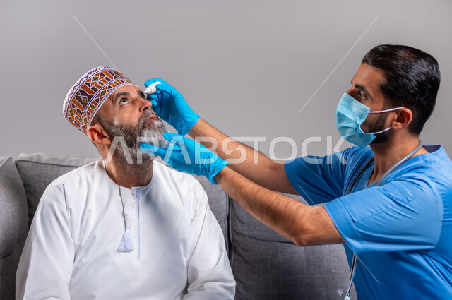 الاهتمام بالأعضاء الحساسة في الجسد, العمل في القطاع الصحي، مفهوم الطب والرعاية الصحية، بورتريه مقرب لممرض عيون مختص يرتدي اليونيفورم و والكمامه والقفازات الطبية يقوم بوضع قطرة عينية لرجل مسن عربي خليجي عماني يلبس الدشداشة والكمه يجلس على الكنبه، خلفية رمادية