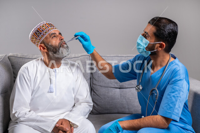 ممرض يلبس الكفوف وكمامة الوقاية يقوم بقياس درجة حرارة الجسم بواسطة جهاز الترمومتر الرقمي، شركات التمريض المنزلي, الحاجة إلى المساعدة الطبية, بورتريه مقرب لرجل مسن عربي خليجي عماني يرتدي الدشداشه والكمه يجلس على الكنبة مصاب بالحمى, خلفية رمادية